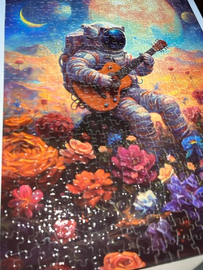 This image captures a completed jigsaw puzzle, presumably located in Santiago, Panama. The puzzle itself depicts a vibrant, fantastical scene. The central subject is an astronaut, clad in a white and blue-striped spacesuit with a reflective helmet visor, positioned in the mid-ground. The astronaut is actively playing an orange electric guitar with a black fretboard, strumming with the right hand and fretting with the left. The astronaut appears seated or floating amidst an abundant field of large, colorful flowers, including prominent orange, red, pink, purple, and blue blossoms. Small, scattered white light specks are visible in the lower foreground. The celestial background features a distinct yellow crescent moon and a larger, reddish-orange planet or moon against a gradient sky that transitions from deep blue at the top to warm orange and yellow hues near the horizon, suggestive of an otherworldly sunset or sunrise. The entire image is rendered in an artistic, illustrative style, with the visible grid lines characteristic of a jigsaw puzzle.