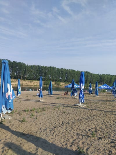 This media file depicts a beach scene on a sunny day with a clear, blue sky with scattered white clouds. The foreground is dominated by sand with a few sparse patches of vegetation. Several closed blue and white beach umbrellas are set up on the sand. In the mid-ground, a calm river flows, with a line of lush green trees forming a dense forest along the opposite bank.
Several people are visible near the riverbank and in the water, suggesting a leisure activity like swimming or sunbathing. The overall atmosphere is relaxed and peaceful, indicative of a typical summer day at the beach. The time of day appears to be late afternoon, given the angle of the shadows cast by the umbrellas.
Visible text on the umbrellas includes "OM" and "este decizia mea". The location context of Vadul lui Voda, Moldova, places this scene by the Dniester River.