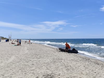 A wide, sandy beach in Yantarny, Russia, is shown on a clear, sunny day. The turquoise ocean with moderate waves stretches to the horizon under a bright blue sky with scattered white clouds. In the foreground, a man wearing an orange vest and dark shorts stands next to a dark inflatable boat with an outboard motor on the wet sand near the waterline. The boat has Russian text printed on its side. In the mid-ground, several people are scattered across the beach, some sunbathing on towels and others walking. Further in the background, a lifeguard tower with a yellow flag stands on a dune. To the left, a white building structure is visible. The overall scene depicts a typical summer day at a coastal resort.