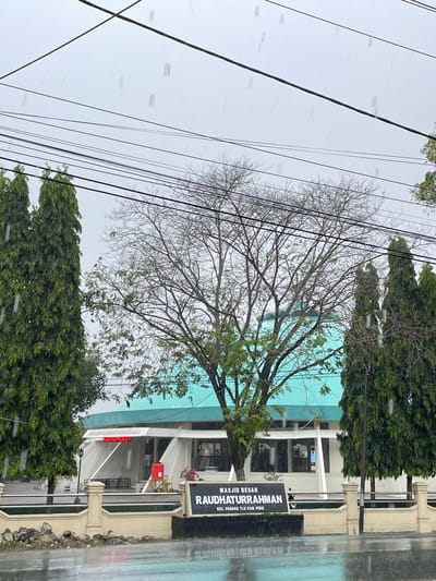The image shows the Masjid Besar Raudhaturrahman in Pasar Paloh, Indonesia, on a rainy day. The mosque has a prominent light blue dome and white architectural features. In front of the mosque, there is a sign displaying "MASJID BESAR RAUDHATURRAHMAN" and "KEC. PADANG TIJI KAB. PIDIE". Tall, dense green trees are on either side of the mosque, and a bare-branched tree stands in front of the dome. The ground in the foreground is wet and reflective, indicating heavy rain. Overhead, power lines crisscross the sky, which is overcast and grey. There are no people visible in the image.