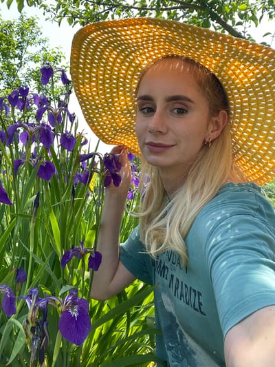 A young woman with blonde hair, wearing a teal t-shirt and a large yellow sun hat, poses for a selfie in a garden filled with purple irises. The woman is positioned in the right half of the frame, with her face turned towards the camera, a slight smile on her lips. Her left hand is visible, resting near the irises. Lush green foliage and the bright sky form the background. The scene suggests a sunny day, likely outdoors. The location is stated to be Knevichi, Russia.