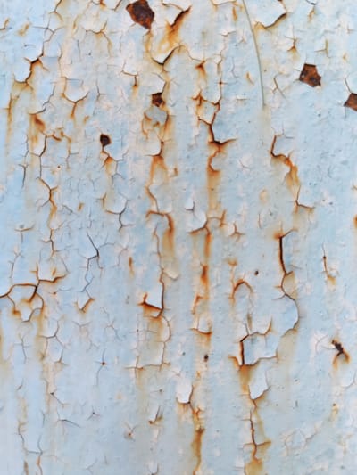The image displays a close-up, abstract view of peeling light blue paint revealing rusty patches underneath. The texture is characterized by numerous fine cracks in the paint, creating a web-like pattern. The rusty areas vary in size and shape, with some appearing as streaks and others as irregular blotches. The lighting suggests an outdoor environment, potentially a weathered surface. Without further information or context linking this image to Borodino, Russia, no specific details about its location within the city can be provided. The image solely focuses on the surface texture and does not contain any people, identifiable objects, or discernible actions.