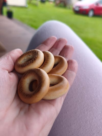 A close-up shot shows a person's open hand holding five small, ring-shaped pastries. The pastries are golden brown and appear to be dry and slightly crisp, with visible indentations or lines on their surfaces. The hand is gently cupping the pastries, with fingers slightly curled. In the background, out of focus, is a blurred expanse of green grass, a paved surface that appears to be a porch or walkway, and a red car parked in the distance. The scene suggests an outdoor setting, possibly a backyard or park, in the city of Brodi, Latvia. The overall lighting indicates it is daytime.