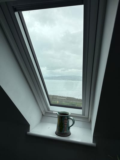 This vertical shot captures an interior view looking out through a window, possibly a skylight or dormer window. The window pane is partially obscured by numerous water droplets, suggesting recent rain or spray, and reveals an overcast, cloudy sky. Beyond the immediate sky, a body of water, likely a loch or sea, stretches towards a distant landmass or coastline, consistent with the Sconser, United Kingdom location.

A light blue/green ceramic mug is prominently displayed on the white windowsill to the right. The mug is decorated with a distinct fish pattern, featuring red and white striped bodies and greenish-yellow fins. It appears to be lying on its side, with its handle positioned upwards.

The window frame is white or a light shade, contrasting sharply with the dark grey or black wall surrounding the window recess. The white windowsill shows some minor cracks or imperfections in the paint at the upper right corner where it meets the wall. The scene is brightly lit by the natural light filtering through the window, while the interior room remains largely in shadow. No people or visible text are present in the image. The overall impression is a quiet, contemplative scene on a cloudy or rainy day by the water.