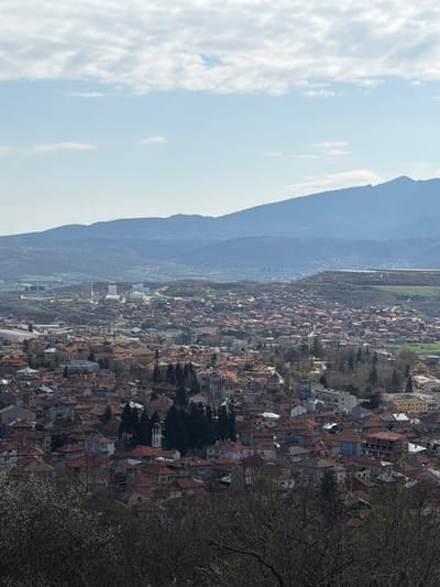 An aerial view depicts the city of Sveti Konstantin, Bulgaria, extending from the base of a mountain range toward the sky on the left. The sky contains scattered clouds. The cityscape consists of dense arrangements of buildings with terracotta roofs. Trees punctuate the urban landscape. In the foreground on the right is foliage.