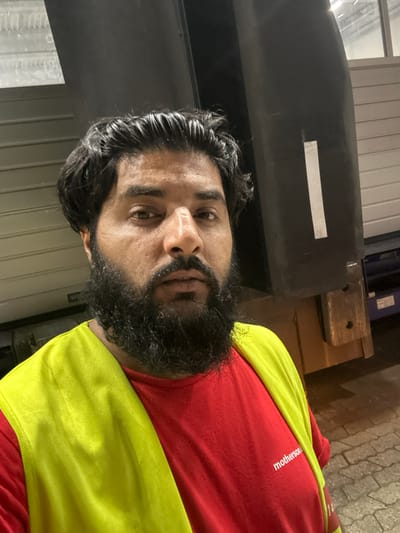 A close-up, slightly angled, selfie-style photograph shows a man with dark, wavy hair and a full, dark beard. He is wearing a red t-shirt with "motherson" printed in white text on the lower right side of the shirt. Over the red t-shirt, he wears a bright, neon yellow safety vest. The man is outdoors at night, evidenced by the lighting. In the background, there is a large, dark industrial structure that appears to be a truck trailer or shipping container, with horizontal paneling visible on its surface. There is also a lighter-colored, ribbed surface to the left of the frame, possibly part of a building or another trailer. The ground appears to be paved with light-colored interlocking bricks. The overall setting suggests an industrial area or logistics hub. The location of Schweitenkirchen, Germany, is not discernible from the visual elements of the image itself, but is provided as context.