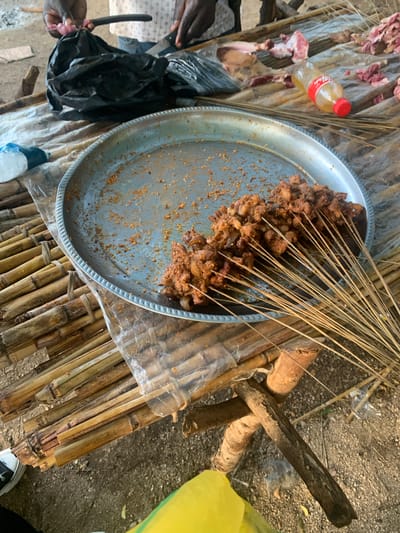 This media file captures a scene with food preparation in Reak, Nigeria. The primary subject is a large metal tray filled with skewers of cooked meat. A bundle of thin wooden skewers is placed on top of the meat, suggesting it has just been prepared or is ready for serving. Surrounding the tray, the setting appears to be outdoors, with a ground covered in sand and small stones. Various objects are scattered around, including raw meat on a plastic sheeting, a red-capped plastic bottle of a beverage, and black plastic bags. There are also bundles of bamboo or similar reeds, possibly used for cooking or as part of the preparation surface. In the background, a bright yellow bag is visible. The overall impression is that of a casual, perhaps informal, food gathering or market setting. The lighting suggests it could be daytime, with natural light casting shadows. There is no visible text in the image. The activity taking place is the preparation or presentation of grilled meat skewers. The focus on the food and the rustic setting evoke a sense of local culinary tradition.