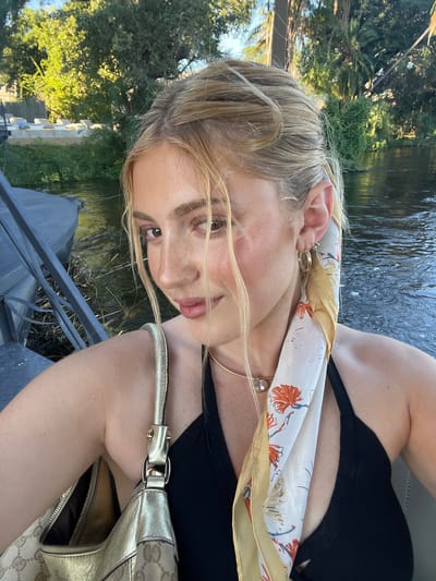 A fair-skinned woman is captured in a close-up, tilted-angle shot. She wears a black tank top and a patterned scarf, with a golden necklace and earrings. The strap of a gold-colored purse is visible. The background consists of greenery and a body of water that could be the Zambezi river. A metal railing can be seen in the foreground. Based on the scenery, it is possible the photograph was taken near the Zambezi river. The precise location within Livingstone, Zambia, cannot be determined from this image alone.