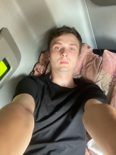 In a close-up, overhead shot, a young Caucasian man with short brown hair and light brown eyes is lying down on a bed with a pink and brown patterned comforter. He is wearing a black t-shirt and looking directly at the camera. His right arm is extended towards the camera. To the left of the man, a grey panel with a bright green insert is visible. The background appears to be the interior of a vehicle, likely a truck cab, based on the grey panel and the confined space. The lighting is dim, creating shadows on the ceiling of the cab. The image does not provide any specific details or clues that would allow for pinpointing a location within Brachterbeek, Netherlands.