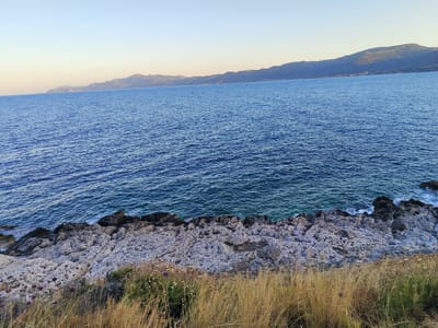 The image depicts a rocky shoreline in Gefyra, Greece. The sea, a deep blue, stretches across the frame, meeting the land at a jagged, light-colored rocky coast. The rocks are uneven and textured, with some darker, more rugged formations closer to the water's edge. In the foreground, dry, golden grass and some flowering plants with purple heads grow sparsely. In the distance, a range of hazy mountains forms the horizon under a pale, clear sky. The scene appears to be captured during daylight, likely late afternoon or early evening, given the soft lighting. There are no people or visible structures in the image.