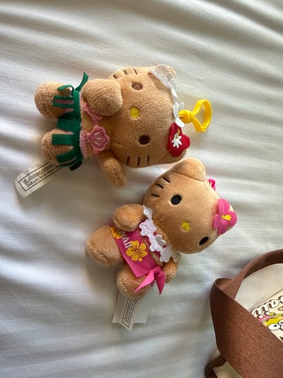 Two plush Hello Kitty dolls are lying on a white, slightly wrinkled surface. One doll wears a green hula skirt, a seashell necklace, and a flower in its hair, with a yellow plastic hook attached to its head. The other doll is dressed in a pink dress with floral accents and a matching flower in its hair. Both dolls have brown fur and are lying next to a brown bag strap. A tag with a warning label is visible near the doll with the hula skirt. The location is unknown.