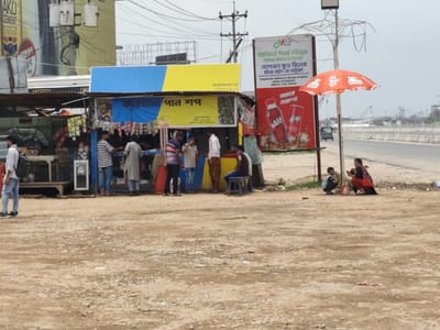 The image captures a roadside scene in Hatikumrul, Bangladesh. A small stall, labeled "Farhana Pan Shop" in Bengali, is the focal point, serving customers. Nearby, a sign advertises "National Food Village Highway Hotel & Restaurant." Several people are gathered around the stall, possibly engaging in transactions or conversations. A woman and child sit on the ground near the food village sign. An orange umbrella shades them. The area is dusty and appears to be along a highway or major road, as evidenced by the visible road and overpass in the background. There is trash on the ground. Electrical wires are overhead. A billboard is also visible in the background.