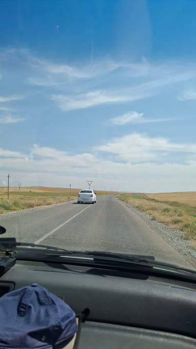 The image shows a view from inside a vehicle looking out onto a paved road in the countryside. The road curves to the right and stretches into the distance, bordered by dry grass and fields. A white car is driving ahead on the road. The sky is blue with scattered white clouds. In the foreground, the dashboard of the vehicle is visible, with a blue cap on the left side. There are utility poles and wires along the side of the road. The location is identified as Құтарыс, Kazakhstan.