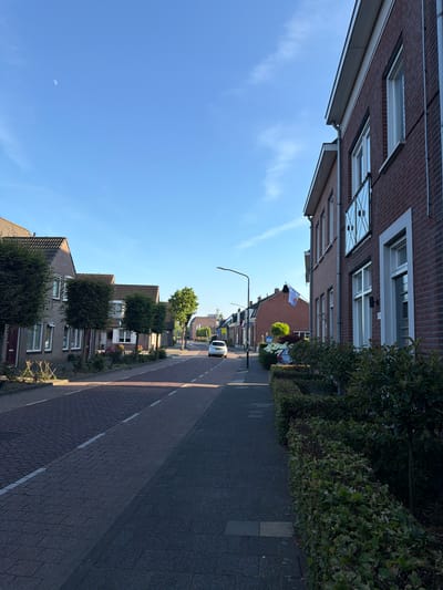 The image depicts a street scene in Boxtel, Netherlands. It is a sunny day with a clear blue sky, and the crescent moon is visible. The perspective is from a low angle, looking up along a street lined with brick houses and green trees. A paved sidewalk runs alongside the street, which is also paved.
On the left side of the street, the side of a brick building is visible, with windows and a gutter system. On the right side, houses with pitched roofs and windows are interspersed with well-maintained greenery. A white car is parked on the street further down, and a streetlamp stands near the middle of the scene. A flag with a white design on a red background is flying from a pole in front of one of the houses. The time of day appears to be late afternoon or early evening, given the low angle of the sun casting long shadows. The overall impression is one of a peaceful, residential street in a European town. There is no visible text in the image.