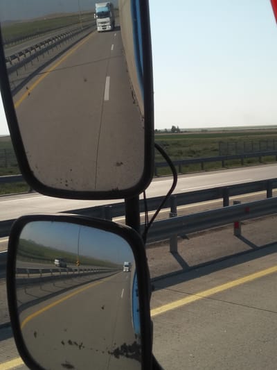 The image shows a view from inside a truck, looking out at the highway through the side mirrors. In the top mirror, a white semi-trailer truck with a blue cab is visible on the road ahead. In the bottom mirror, a highway is reflected, showing a white semi-trailer truck and a white car driving in the same direction. The highway has a yellow line and white lane markings. The surrounding environment is a flat, rural landscape with green fields and a clear blue sky. There are guardrails along the side of the highway. The image is taken during daylight. The location is specified as the city of Баймырза, Kazakhstan, on a highway.