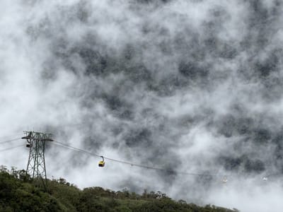 This media file captures a dramatic scene of a cable car system traversing a misty, mountainous landscape. The primary subjects are the cable car pods, with one prominent yellow pod hanging from a steel cable on the right side of the frame, and another partially visible yellow pod closer to a large green support tower on the left. A third white pod is also visible in the distance on the right. The support tower, a tall green metal structure, stands prominently on the left, holding up the cables that stretch across the frame.

The setting is a dense, green forest that covers the steep slope of a mountain. The trees are lush and dark, providing a stark contrast to the swirling white and gray clouds that partially obscure the mountain peaks and the cable car system. The weather is clearly misty or foggy, creating an ethereal and somewhat mysterious atmosphere. The low visibility suggests it's likely a cloudy or damp day.

The activity depicted is the transportation of passengers via the cable car, though no people are clearly visible within the pods. The overall mood evoked is one of awe and perhaps a touch of isolation, given the vastness of the landscape and the obscuring mist. The image conveys the scale of the operation and its integration into the natural environment. There is no visible text in the media file.
