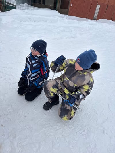 The image depicts an outdoor, snow-covered scene in Zaosinovo, Russia, featuring two young males, likely children, dressed in winter attire. The ground is uniformly covered in white snow with visible boot prints. In the background, a green metal fence or grate is partially obscured by snowdrifts on the left, while a reddish-brown wooden building with a vertical plank door and a dark numerical sign (possibly "30") is visible on the right.
On the left, one child is kneeling on the snow. He wears a dark blue knit beanie, blue-framed eyeglasses, a multi-colored winter jacket with blue, black, white, and red geometric patterns, black winter pants, black gloves, and black boots. His head is turned slightly to the right, and he exhibits a slight smile.
On the right, a second child is in a crouching or kneeling position. He wears a light blue knit beanie adorned with a red star emblem, a camouflage-patterned winter overall suit in shades of green, brown, black, and yellow, dark blue gloves, and black winter boots. He is actively holding two clear icicles: one long, vertical icicle is held by his right gloved hand, extending downwards, and a shorter, horizontal icicle is held by his left gloved hand near his left knee. He is looking downwards and to his left, seemingly focused on the icicles or the snow.