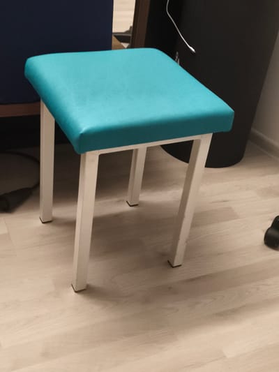 The image shows a teal upholstered stool with white metal legs on a light-colored wooden floor. The stool is square and appears to be in a room with a dark cylindrical object and a dark blue chair in the background. The lighting is dim, casting shadows on the floor. No specific location in Troisdorf, Germany can be determined from the image.
