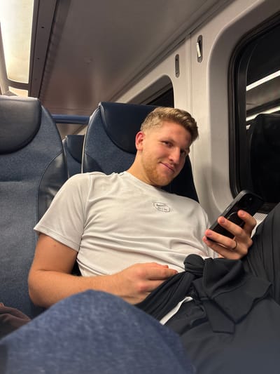 A male subject is seated on a passenger train in Edison, United States. The individual, appearing to be in his late 20s to early 30s, has short light brown hair and facial stubble. He is wearing a light grey short-sleeved t-shirt with a "Nike Air" logo on the chest. He wears a dark ring on the fourth finger of his left hand. In his left hand, he holds a black smartphone, which features a prominent rear camera module and a MagSafe ring. A crumpled black garment is resting on his lap. He is situated in a blue-upholstered train seat with a dark headrest, looking directly at the camera with a slight smile. Another identical train seat is visible to his left. The train interior exhibits white ceiling panels with integrated lighting and a window on the right showing a dark exterior.