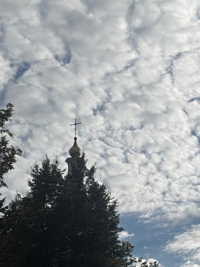 The image shows a close-up view of the top of a church spire, adorned with a cross, peeking through dense foliage of trees. The spire has a golden dome and a dark gray cross. The background is a sky filled with white, textured clouds interspersed with patches of blue sky. The setting appears to be outdoors, possibly in a park or a churchyard, in the city of Pereiaslav, Ukraine. The lighting suggests it is daytime, with the sun likely overhead, casting soft light on the clouds. There are no people or visible actions depicted in the image.