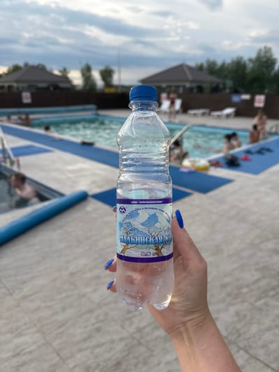 In Yuzhnyye Koryaki, Russia, a hand with blue nail polish holds a partially frosted plastic bottle of water in the foreground. The bottle has a label with a winter landscape and Cyrillic text. In the background, a swimming pool area is visible, with several people in and around the water. Loungers and umbrellas are arranged around the pool, and buildings with pitched roofs are situated in the distance under a cloudy sky. The ground surrounding the pool is paved with light-colored tiles.