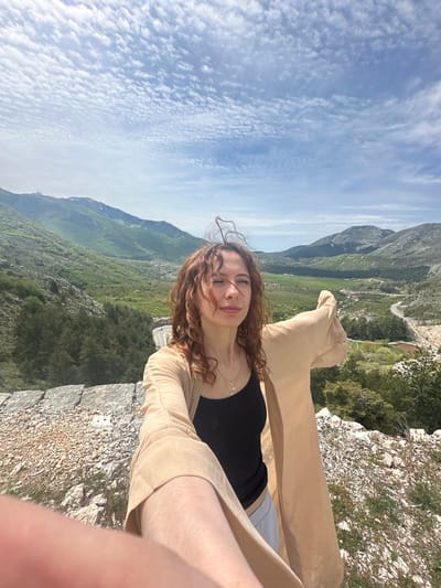 A woman with curly, reddish-brown hair and fair skin is standing on a stone ledge, smiling with her eyes closed. She is wearing a black top and a light tan, open-front cardigan. The background consists of a mountainous landscape covered in greenery and trees. The sky above is blue with scattered white clouds. The location is likely Bukovica, Montenegro, considering the characteristic terrain and vegetation.