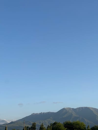 This is a vertical, medium-shot, outdoor photograph of a mountainous landscape under a clear blue sky, taken in the village of Sarymoldayev in Kazakhstan. The upper two-thirds of the image is filled with the vast expanse of the sky, transitioning from a lighter hue at the horizon to a deeper blue at the top. Faint, wispy clouds are visible scattered across the sky. The lower third of the image features the silhouette of a mountain range with jagged peaks. Rolling green hills lead up to the mountains. In the foreground, a line of dark green trees forms a dense border. Several utility poles and wires are visible across the landscape, indicating human infrastructure. The lighting suggests it is daytime, possibly late afternoon or early morning, given the soft light on the mountains. There are no people or animals visible in the scene.