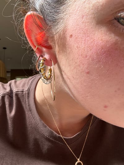 An extreme close-up features the side of a person's face, highlighting their left ear adorned with multiple earrings. There's a collection of hoops, spirals, and a larger earring with interwoven gold and silver rings. A delicate gold necklace with a circular pendant drapes across the upper chest and shoulder. The person's skin exhibits freckles and redness, contrasting with graying hair near the ear. The focus is primarily on the ear and its jewelry.