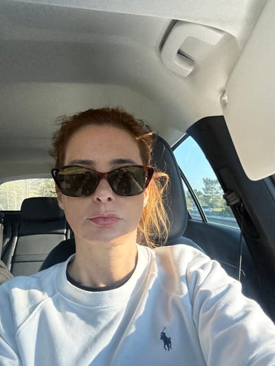 The image captures a female individual seated in the front passenger seat of a vehicle during daylight. She has reddish-brown hair, is wearing dark, cat-eye style sunglasses, and a white crew-neck sweatshirt featuring a navy blue Ralph Lauren polo pony emblem on the left chest. A dark undershirt is visible at the neck. Sunlight illuminates the right side of her face and body.

The vehicle's interior includes a light-colored headliner, a visible grab handle above the passenger door, and a partially lowered light-colored sun visor. Dark headrests and seat upholstery are present in the rear. Through the front passenger window, a clear blue sky is visible above a roadside landscape featuring a barrier and trees. A light-colored vehicle is discernible on a road in the distant background. The specific location within Vagos, Portugal, is not discernible from the visual information provided beyond the general context of being in a moving or parked vehicle on or near a road.