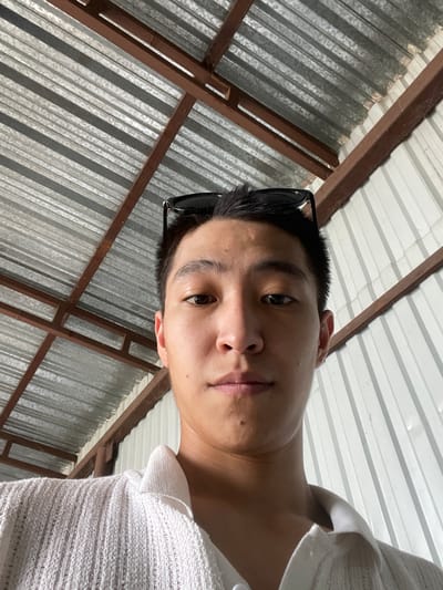 The image shows a close-up, low-angle selfie of a young man wearing a white collared shirt. He has sunglasses perched on his head. The background consists of a corrugated metal roof with visible brown metal supports, and a white corrugated metal wall. The scene appears to be outdoors, possibly under a covered structure. The lighting suggests it is daytime. The location is specified as Кенен Әзірбаев ауылы, Kazakhstan.