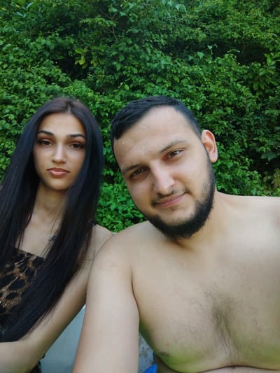 A young adult couple is pictured outdoors in front of a dense green hedge. The female on the left has long, dark, straight hair and is wearing a leopard print top. She has a neutral expression and is looking towards the camera. The male on the right has short dark hair and a full beard. He is shirtless and appears to be smiling at the camera. The scene suggests they are in a backyard or park area, possibly near Herford, Germany, given the greenery.