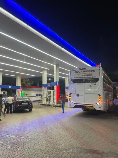 At a brightly lit petrol station in Kajheri, India, a large white bus with "INDO CANADIAN EXPRESS" branding is parked. The bus has illuminated taillights and a license plate reading "AR 11L 1021". A black sedan with license plate "CH01CA2413" is positioned near a petrol pump. Several people are visible, including a man in a red shirt walking away from the camera, and others gathered around the sedan. The environment is characterized by modern petrol pumps with white canopies illuminated by bright white and blue LED lights, and a patterned brick-paved ground. A tall electricity pylon is visible in the dark sky in the background.