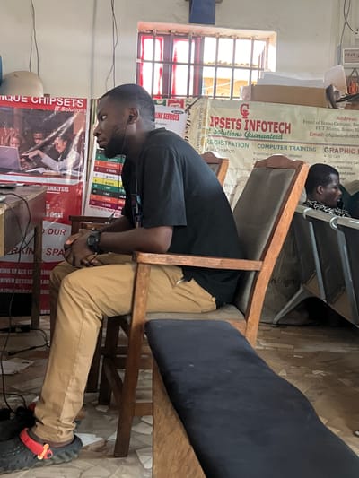 A man sits on a wooden chair, his head bowed forward, looking at something on a desk. He wears a black shirt, khaki pants, and black sandals with red accents. His hands are clasped in his lap and a watch is on his wrist. The background shows a room with promotional posters for "Chipsets Infotech IT Solutions," detailing computer training and graphic design services, with an address indicating the location is near the Bosso Campus of FUT Minna in Bosso, Niger State, Nigeria. Another person is seen lying down on a rack in the back. The room appears to be in a state of clutter.