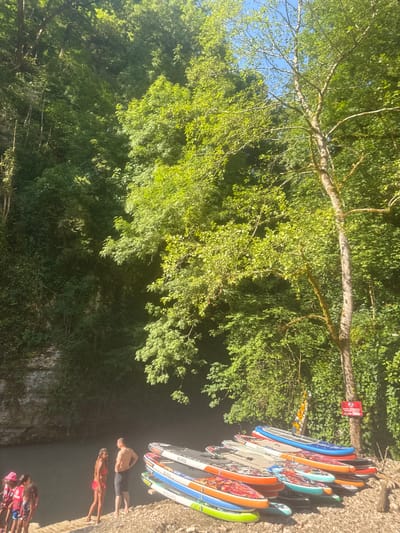 In Khsyrkhu, Abkhazia, a group of people are gathered near a body of water. In the foreground, several stand-up paddleboards are neatly stacked on a pebbly shore. A man and a woman are conversing on the shore, with the man wearing dark shorts and the woman in a pink bikini. To the left, a group of younger individuals, some wearing life vests, are standing on a wooden platform. The background is dominated by lush, green vegetation covering steep rock faces and a dense forest canopy under a clear blue sky.