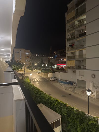 The image depicts a street scene at night in Fuengirola, Spain. In the foreground on the left, a white arched balcony railing and part of a building are visible, suggesting a viewpoint from a hotel or apartment. To the right, a multi-story white apartment building features balconies with railings, air conditioning units, and yellow awnings. Clothes are hanging on a line on one of the balconies.
The street is paved and illuminated by streetlights. A white car is parked on the side of the road. A blue dumpster filled with debris is also present on the side of the street, near some construction fencing. Across the street, there is a building with a prominent red sign that reads "SUPERMERCADO". Further in the background, other buildings and lights suggest a residential area on a hillside. A well-maintained hedge runs along the side of the street, separating the road from the balcony in the foreground. A black lamppost with an illuminated lantern stands on the sidewalk.