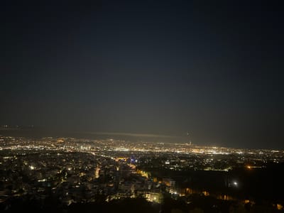 The image is a nighttime aerial view of a city, likely Athens, Greece, taken from a high vantage point. The foreground is dominated by dark silhouettes of trees and possibly some buildings on a hillside. Below, the city stretches out, a vast expanse of twinkling lights representing buildings, streets, and illuminated landmarks. The sky above is a dark, deep blue, with a few faint stars visible. In the distance, faint atmospheric haze can be seen, obscuring some of the city lights. The overall impression is one of a sprawling urban landscape at night, with the lights creating a warm glow against the dark sky. The location is identified as Chaidari, Greece, a municipality in the northwestern part of the Athens metropolitan area.