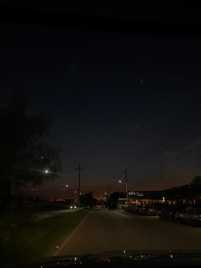 The image depicts a nighttime scene from the interior of a moving vehicle on a paved road in Xcanatún, Mexico. The sky is dark blue-black with a discernible orange-red glow on the horizon.
On the left side of the road, there is a grassy shoulder or median with blurred dark foliage, indicative of motion. Multiple utility poles with power lines extend alongside the road. The bright headlights of at least one oncoming vehicle are visible in the distance.
On the right side, a commercial building with the illuminated sign "RockSport Plazas" is visible. A parking lot in front of this building contains numerous parked vehicles. Additional utility poles and power lines run parallel to the road on this side. Streetlights are active along both sides of the road, casting illumination. Reflections from the vehicle's interior are visible on the windshield in the foreground. No discernable individuals are visible in the scene.