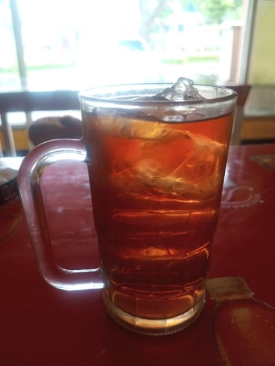 A tall glass mug filled with iced tea sits on a red patterned surface. The glass is clear, revealing the amber liquid and ice cubes. A transparent handle is attached to the side. A window is in the background, emitting bright light, with glimpses of greenery and a building. The table surface has a faint bottle-shaped design. The mug is the focal point.