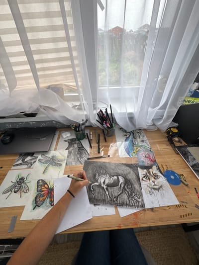 An aerial shot captures a desk in what is presumed to be Oliki, Russia, laden with various artistic creations and supplies. The desk is bathed in natural light streaming through a large window, adorned with sheer white curtains that gently diffuse the sunlight. Spread across the wooden surface are several detailed drawings, including a dragonfly, a fly, a butterfly, a crow, a horse, and a cat. A woman's arm, with a black bracelet on her wrist, is visible as she actively draws on a sheet of paper, holding a pen in her right hand. Nearby, a collection of pens and markers stand upright in a green glass container, along with an open laptop and a computer mouse. The background reveals a glimpse of the outdoors, with greenery, houses, and a fence visible through the window, hinting at a residential setting. The scene suggests a dedicated artist in the midst of their creative process.