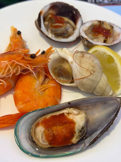 A close-up, high-angle view depicts a selection of cooked seafood presented on a white ceramic plate. In the upper-left quadrant, two bright orange, whole prawns or shrimp are visible, featuring intact heads, antennae, and segmented bodies. To the upper-right, two open bivalve shells, identifiable as clams or cockles, each contain mollusk meat partially covered with a reddish-orange sauce. Adjacent to these, a pale beige, spiraled whelk shell houses a mollusk, with a thin red filament draped across its surface. A single yellow lemon wedge rests next to the whelk and clams. In the lower-right quadrant, a large, open mussel shell, displaying an iridescent blue-green rim, reveals its interior meat, also topped with a reddish-orange sauce. The background is out of focus, with faint, blurred numeric text partially visible in the upper region. No individuals are present. The scene is located in Audun-le-Tiche, France, depicting a culinary presentation.