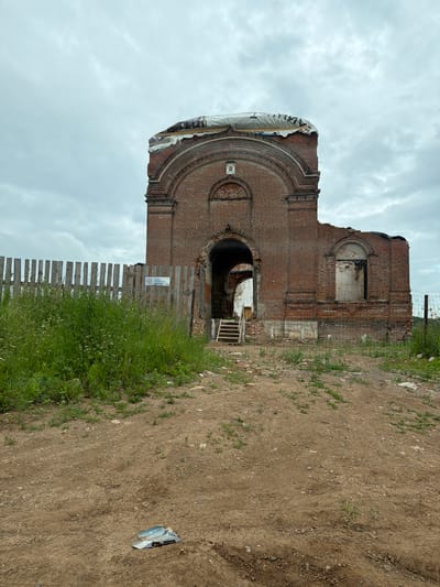 The image depicts an exterior view of a decaying brick structure, possibly a church or bell tower, in the city of Yug, Russia. The building is in a state of ruin, with parts of the roof missing and the walls crumbling. A temporary covering made of plastic sheeting is visible on the upper section of the structure. A wooden fence, partially covered in tall grass, encloses the area. A metal fence is also present, separating the foreground from the structure. A set of wooden stairs leads into an arched opening of the building. The ground in the foreground is dirt and gravel, with scattered debris, including a flattened piece of metal or paper. The sky is overcast with clouds.