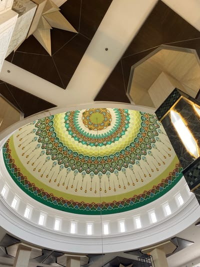The image shows the interior of a building, likely a mosque, in Aluekambuk, Indonesia. The main focus is a domed ceiling adorned with intricate, colorful Islamic geometric patterns and calligraphy. The dome is painted in shades of cream, green, yellow, and brown, with repeating motifs that radiate outwards from a central point. Below the dome, a series of small rectangular windows are visible, set into a white curved architectural element. The surrounding ceiling is a mix of white and dark wood panels, with some angular, multi-faceted wooden structures jutting out. To the right, a tall, dark green, marble-clad structure with a lit interior section, possibly a decorative lamp or architectural feature, is partially visible. The scene suggests a grand and ornate interior space, bathed in ambient light. There are no people or animals visible in the image.