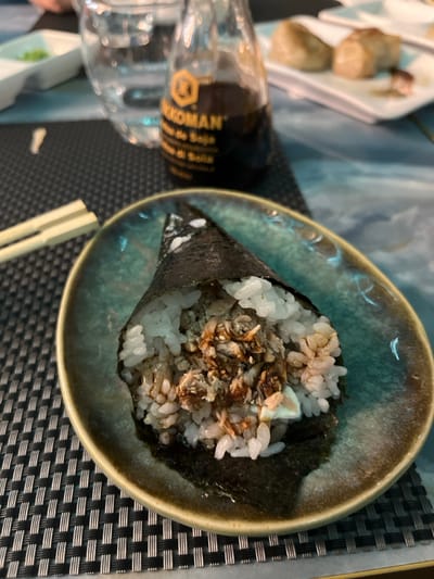 The image displays a close-up view of a Japanese handroll (temaki) presented in a distinctive teal and brown ceramic bowl. The handroll is cone-shaped, composed of white sushi rice and a filling that includes what appears to be shredded protein (possibly fish), a light-colored creamy sauce, and is topped with small, darker crispy bits, likely fried onions or flakes, and a dark drizzle, possibly a sweet soy glaze. The ingredients are encased in a dark green sheet of nori seaweed.

The bowl is set on a dark, woven placemat. To the left, a glass bottle of Kikkoman soy sauce is partially visible and out of focus, with the brand name "KIKKOMAN" and "SALSA DE SOJA" clearly printed on its label. A pair of light-colored chopsticks rests on the placemat to the lower right of the bowl. In the blurred background, other items on a white tray suggest a broader meal setting, likely appetizers. The scene takes place indoors, indicative of a restaurant, and captures a moment during a meal, potentially in Basiglio, Italy. The lighting suggests artificial indoor light, making the time of day ambiguous but often associated with evening dining.