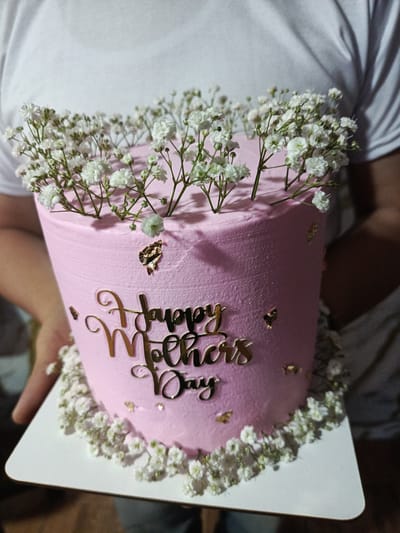 A close-up shot shows a pink frosted cake held by a person or people wearing a white t-shirt. The cake is decorated with baby's breath flowers on top and around the base. A gold-colored "Happy Mother's Day" cutout is adhered to the front of the cake, along with small, scattered gold flakes. The cake sits on a white square cake board. The location is presumed to be Quevedo, Ecuador.