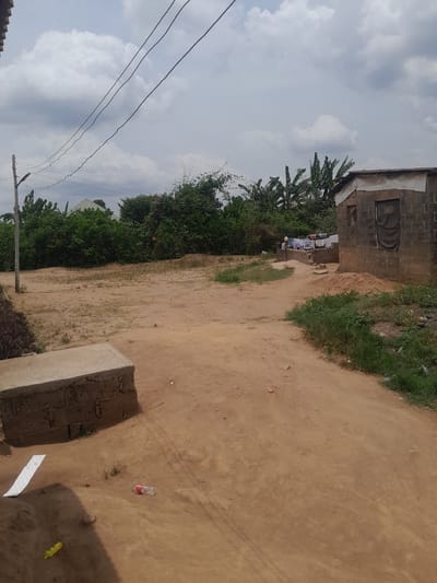 The scene, set in Ifo, Nigeria, depicts an unpaved reddish-brown dirt landscape under an overcast sky with scattered grey clouds. In the lower-left foreground, a low concrete block structure is visible. Adjacent to it on the dirt are scattered items: a red plastic bottle, a small red plastic cap, a piece of white paper, and a yellow plastic object. A slender utility pole stands on the mid-left, from which three dark power lines extend diagonally across the upper frame. To the right, a single-story building constructed of grey concrete blocks features a light-colored material along its roofline and a dark fabric covering a rectangular opening on its facade. A smaller, open window is present to the left of this covered opening. Behind this building, a lower concrete platform supports clothes drying on a line. Dense green vegetation, including bushes and banana trees, occupies the background and mid-ground left. No human figures are visible.