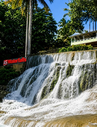 The image displays a cascading waterfall with lush green vegetation surrounding it. The water flows over moss-covered rocks, creating a dynamic and frothy effect at the base. In the background, palm trees and other dense foliage are visible, suggesting a tropical or subtropical environment. A building with a light green railing and a blue roof is perched on a ledge above the waterfall, with a white railing and decorative elements. A sign with red lettering that reads "AS Waterfalls" is mounted on a concrete structure to the left of the waterfall. The sky is clear and blue. The location is identified as the city of Silaiyajae, Indonesia.