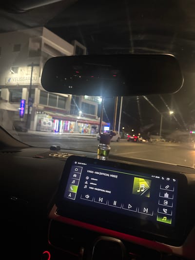The image shows the interior of a car at night, viewed from the passenger seat. The dashboard features a modern infotainment system displaying song information for "V:RGO - HIM [OFFICIAL VIDEO]" with playback controls. The display also shows the time as 23:24 and a temperature of 28.5°C. A car air freshener hangs from the rearview mirror. Outside the car, storefronts with illuminated signage are visible, with one sign displaying "VELISSARIDOU". The street appears wet, suggesting recent rain. Cars and streetlights are visible in the distance. The overall impression is a nighttime drive through a commercial area, possibly in Yeroskipou, Cyprus, given the signage. There are no people clearly visible.