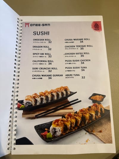The image displays an open menu from a restaurant named "ONEE-SAN", featuring a "SUSHI" section. The menu page includes a "HALAL" certification logo in the top left corner, indicating its compliance for Muslim consumers. Various sushi items are listed with their names in both English and Japanese, followed by their prices. Examples include ONEESAN ROLL (32), DRAGON ROLL (32), SPICY EBI ROLL (32), CALIFORNIA ROLL (34), CHUKA WAKAME ROLL (34), CHICKEN TERIYAKI ROLL (35), CHICKEN KATSU ROLL (35), PIZZA SUSHI CHICKEN (34), DORI CRUNCHY ROLL (32), PIZZA SUSHI TUNA (34), CHUKA WAKAME GUNKAN (34), and ABURI TUNA (32).
Prominently featured on the right side of the page are two platters of sushi rolls, likely for illustrative purposes. These rolls appear to be deep-fried or topped with crispy elements, suggesting a specialty or fusion style. A small dish containing a crispy topping is visible above one of the platters. A pair of black chopsticks rests on a wooden block between the two sushi presentations. The menu itself has a silver spiral binding, indicating it is part of a larger booklet. The setting is likely an indoor environment, such as a restaurant or cafe in Kecamatan Cisaat, Indonesia, given the context. The image captures a detailed look at the food offerings from this particular establishment.
