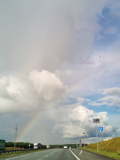 The image depicts a highway scene under a sky with a prominent rainbow arching across dramatic clouds. The highway is asphalt, with clear lane markings. On the left side of the road, there are guardrails and a line of trees in the distance, with a few trucks on the highway. On the right side, there's a grassy embankment with a gas price sign displaying "ДТ ЕВРО 69.50" and a road sign indicating a two-way traffic direction. A few cars are visible on the road. The overall atmosphere is that of a journey through a landscape marked by both natural beauty and infrastructure. The location is identified as Russkie Kirmeni, Russia.