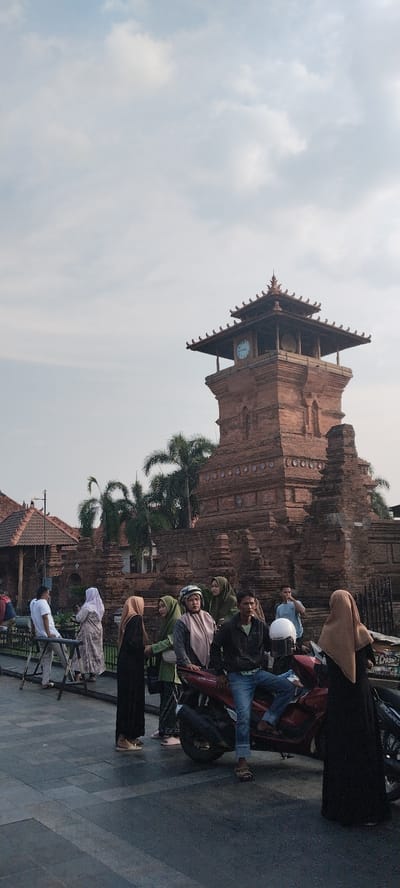 The image captures a scene in the city of Kauman, Indonesia, featuring the iconic Menara Kudus Mosque complex. In the foreground, a group of people are gathered, some standing and others seated on a red motorcycle. The individuals are dressed in a mix of casual and traditional attire, with several women wearing hijabs. The focal point of the background is the striking Menara Kudus Mosque tower, constructed from red brick with a tiered, pagoda-like roof and a prominent clock face. Palm trees are visible behind the tower, adding to the tropical ambiance. The sky is overcast with soft clouds. The scene appears to be a public gathering area, possibly a tourist spot or a place of community interaction.